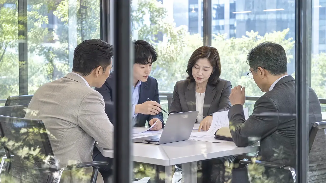 브랜드 영상 장면: 유리벽 너머 회의실에서 네 사람이 서류와 노트북을 보며 논의하는 모습